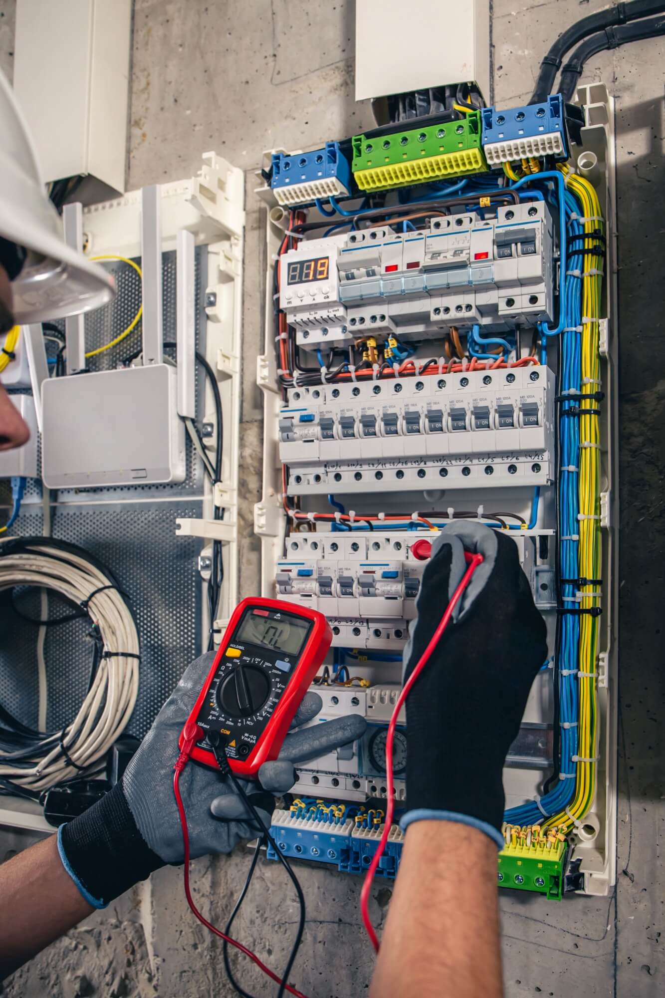 En elektriker med hjälm och handskar använder en multimeter för att mäta spänningen i en elcentral under installation och felsökning.